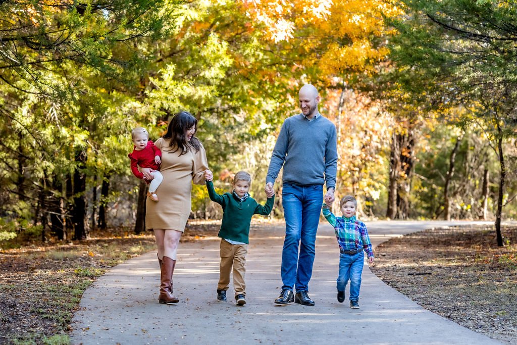 Family of five, mom, dad and three children under the age of 5 holding hands, walking on a sidewalk path at Arbor Creek during a family photo session.