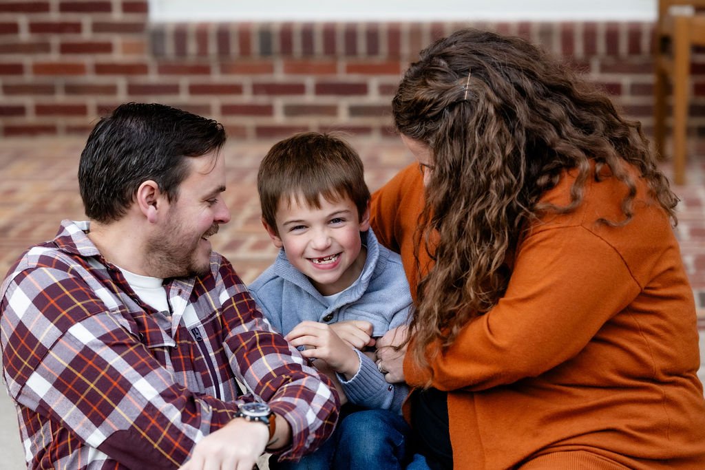 Smiling family of three enjoying a cozy moment during a fall family photo session at Texas Woman's University in Denton, Texas, with warm autumn tones and a brick backdrop creating a relaxed and loving atmosphere