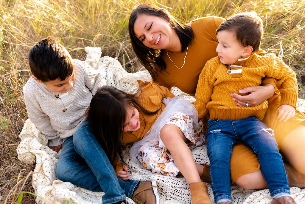 Mom with two boys and one girl sitting on a blanket in a grassy area tickling each other for a photo session.