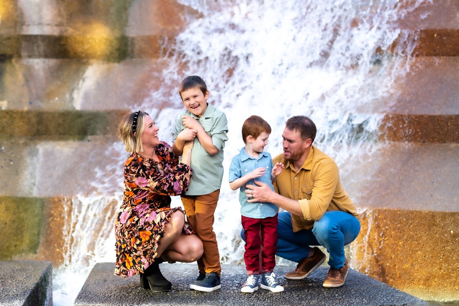 Dad, Mom and two sons aobut 5 years old and 10 years old playfully engaging with each other in front of waterfalling at the Fort Worth Water Gardens in Fort Worth for a family portrait session.