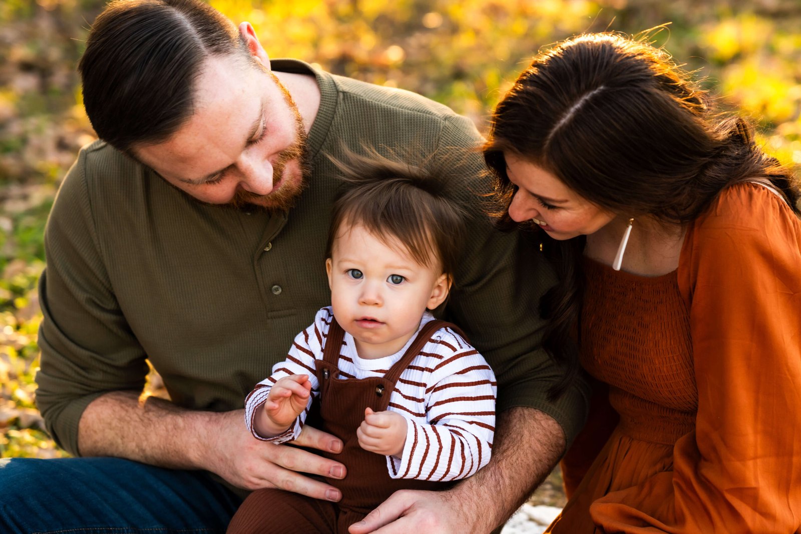 Family of three sitting on the ground at Green Acres Farm Memorial Park in Flower Mound, Texas looking at their baby during a photo session.