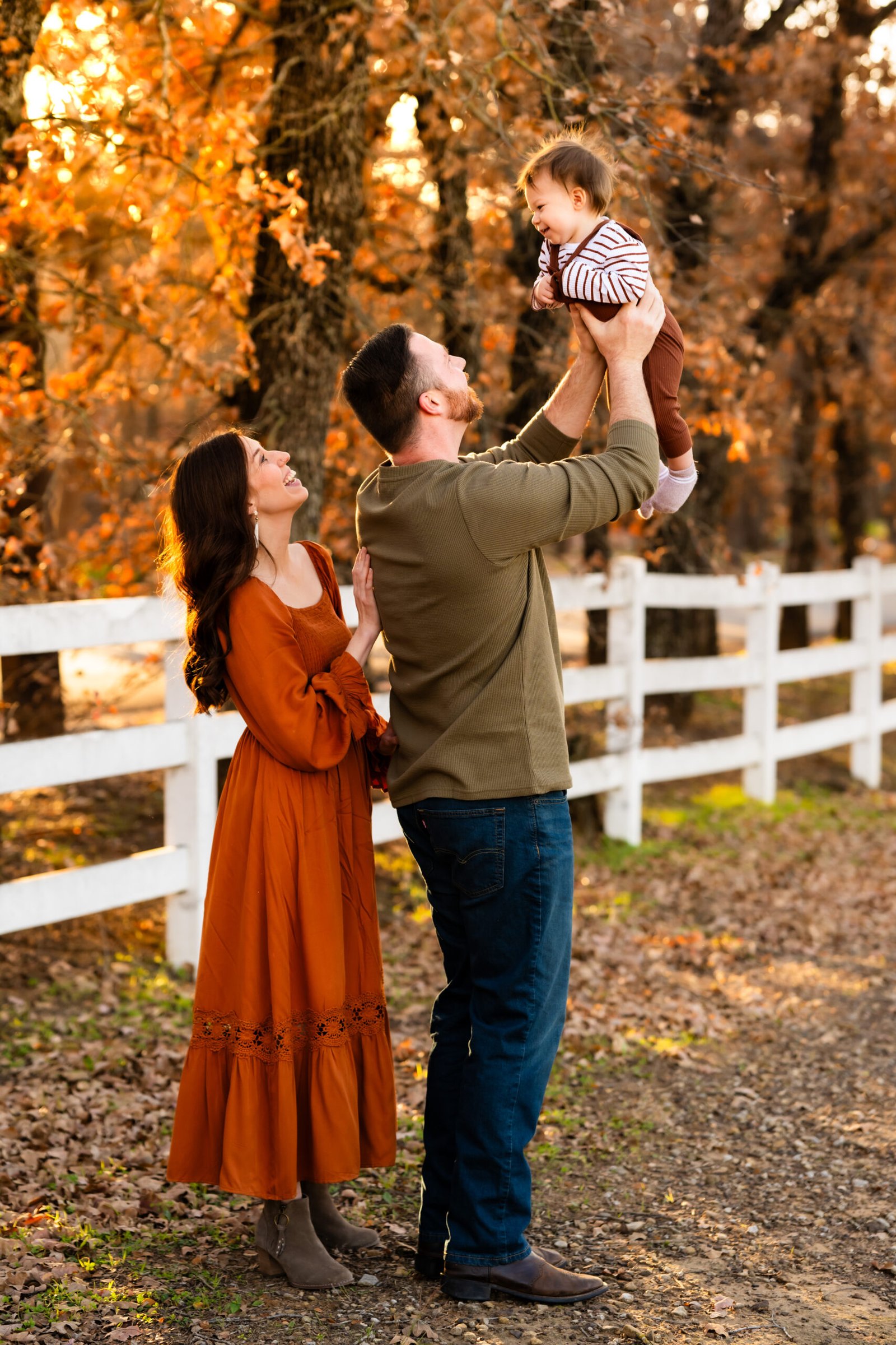 Mom in a rust colored dress standing by Dad in a green shirt tossing their one year old boy in rust colored overalls in front of a white fence with trees playfully looking at each other for a photo session at Green Acres Farm Memorial Park in Flower Mound, Texas.