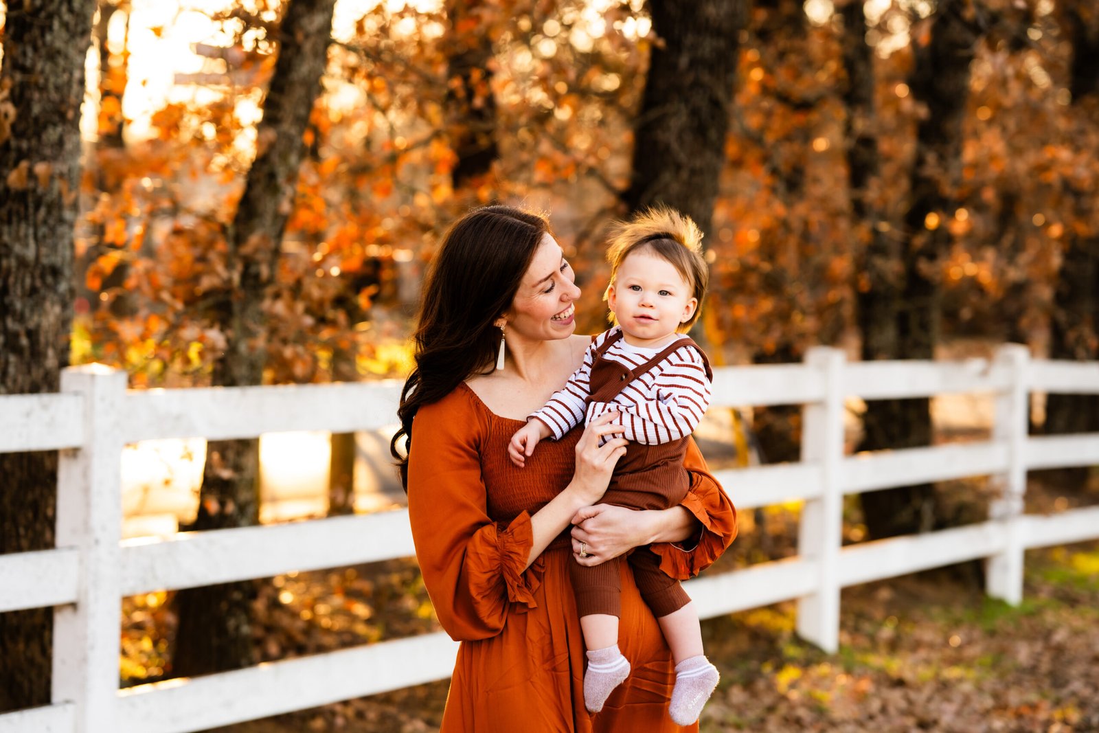 Mom in a rust colored dress holding a one year old boy in rust colored overalls looking at him for a photo session at Green Acres Farm Memorial Park in Flower Mound, Texas.
