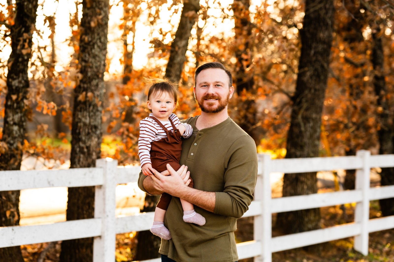 Dad in a green shirt holding a one year old boy in rust colored overalls looking at him for a photo session at Green Acres Farm Memorial Park in Flower Mound, Texas.
