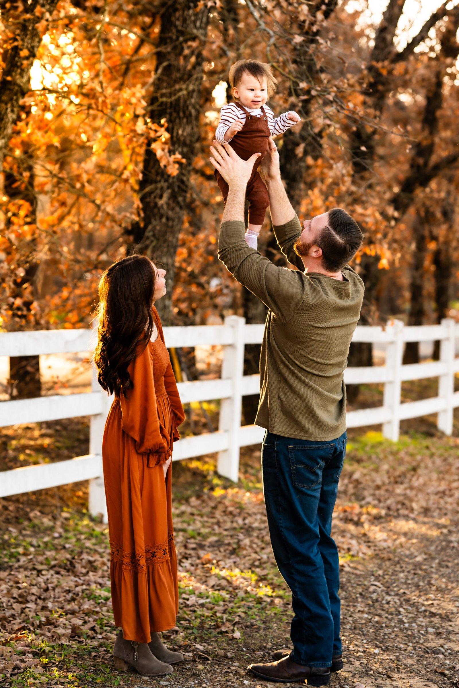 Mom in a rust colored dress standing by Dad in a green shirt tossing their one year old boy in rust colored overalls in front of a white fence with trees playfully looking at each other for a photo session at Green Acres Farm Memorial Park in Flower Mound, Texas.