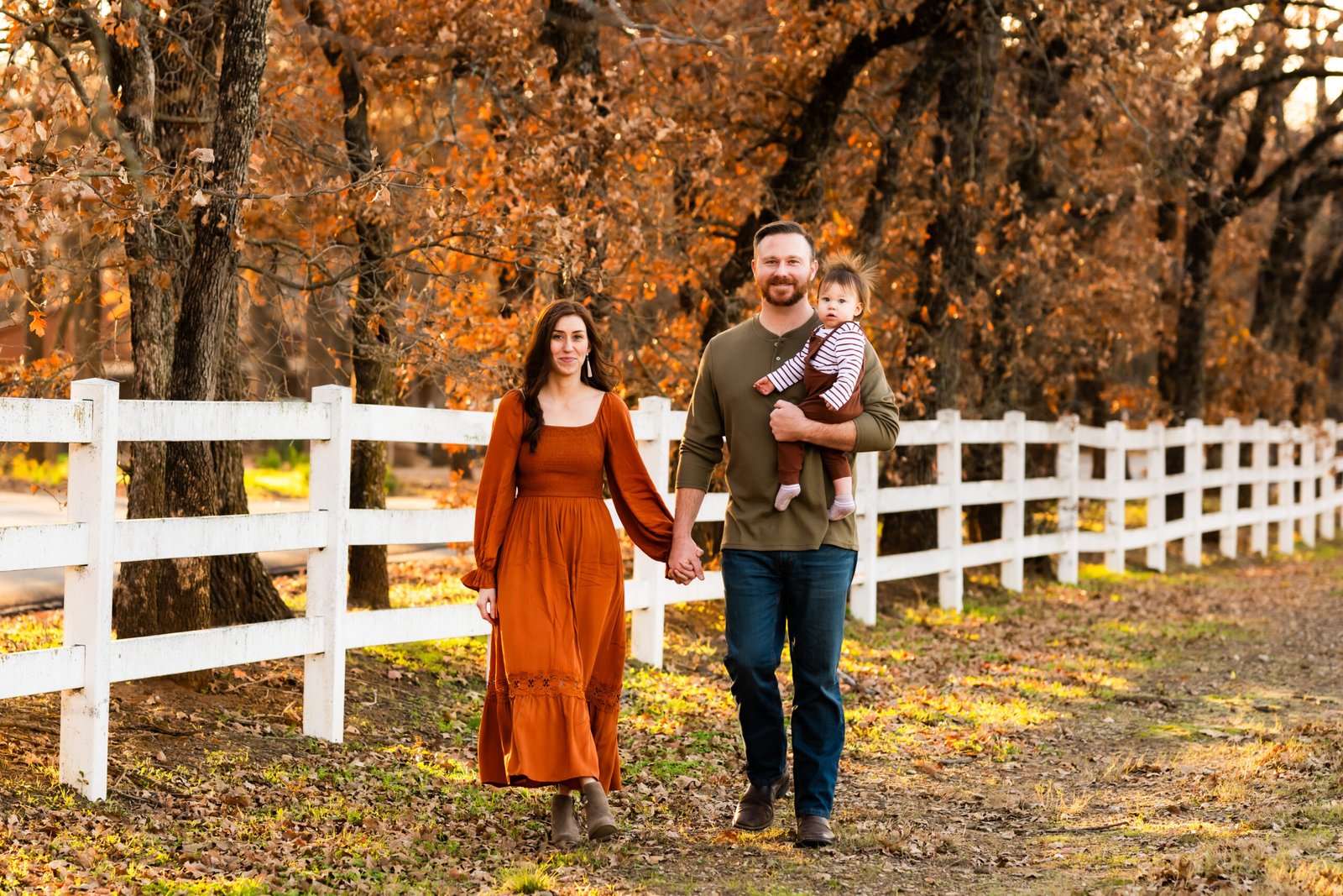 Mom in rust colored long dress holding her one year old boy in rust colored overalls walking by Dad in a green shirt in front of a white fence with fall colored trees for a photo session at Green Acres Farm Memorial Park in Flower Mound, Texas.
