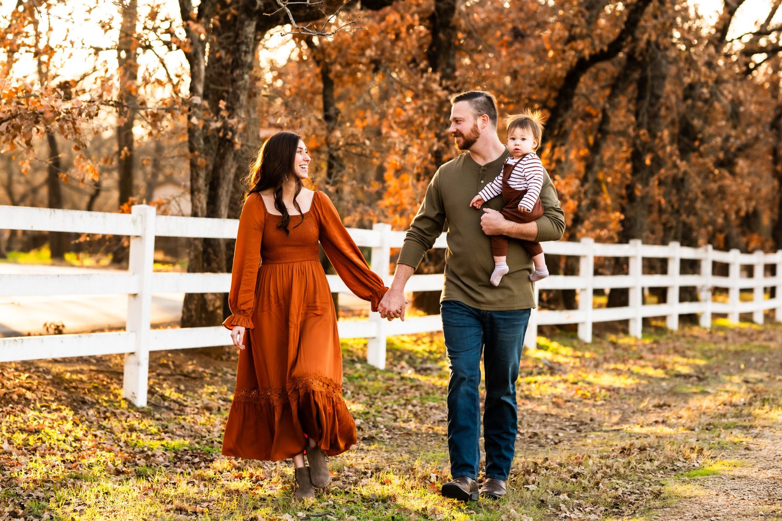 Mom in rust colored long dress holding her one year old boy in rust colored overalls walking by Dad in a green shirt in front of a white fence with fall colored trees for a photo session at Green Acres Farm Memorial Park in Flower Mound, Texas.