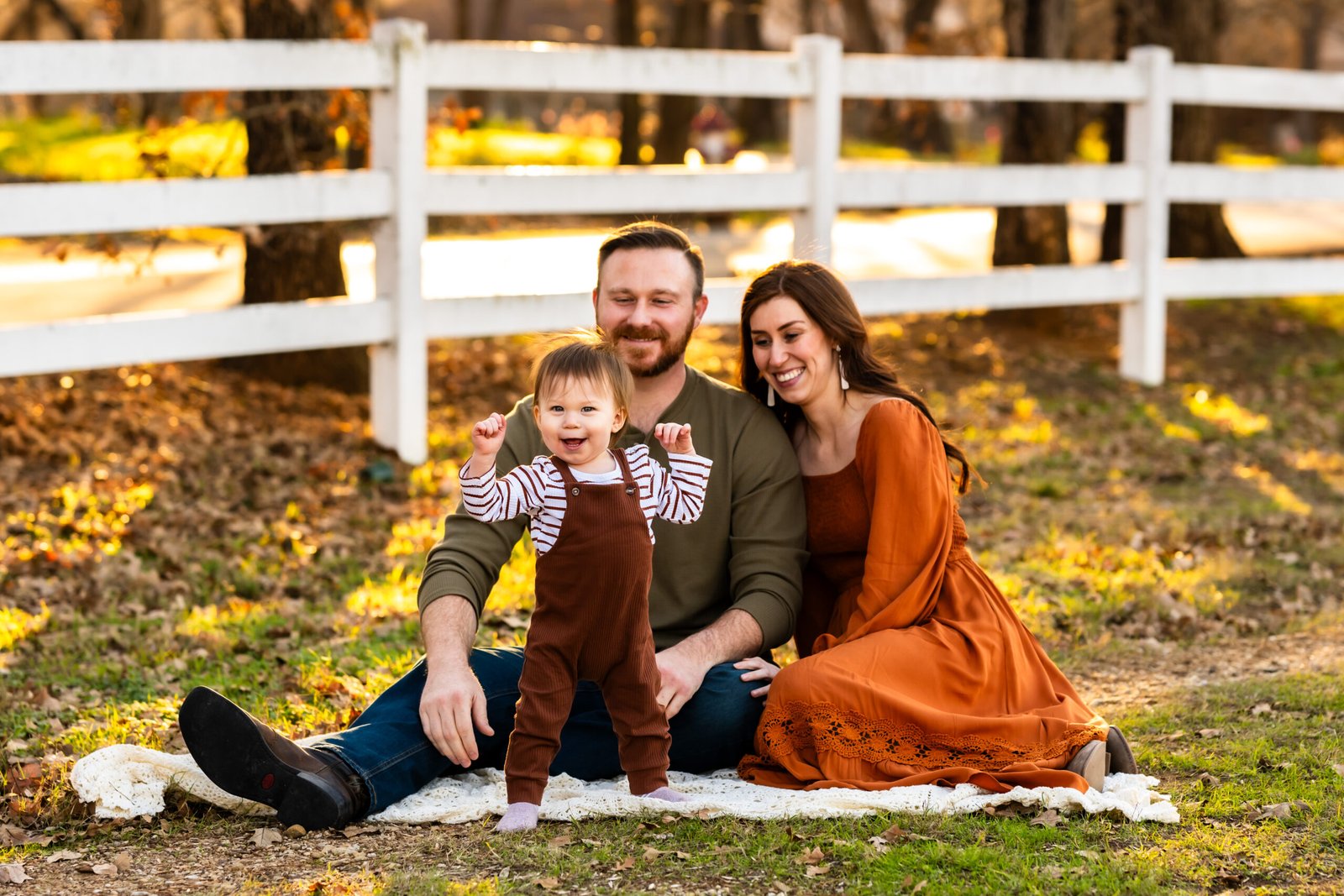 Mom and dad sitting on a blanket with their one year old taking steps away from them at Green Acres Farm Memorial Park posed for a family portrait in December in Flower Mound, Texas.