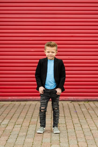 7-year-old boy wearing ripped jeans, a light blue t-shirt, a black blazer, and black-and-white Vans sneakers posing confidently in front of a bold red metal garage door in downtown Roanoke, Texas.