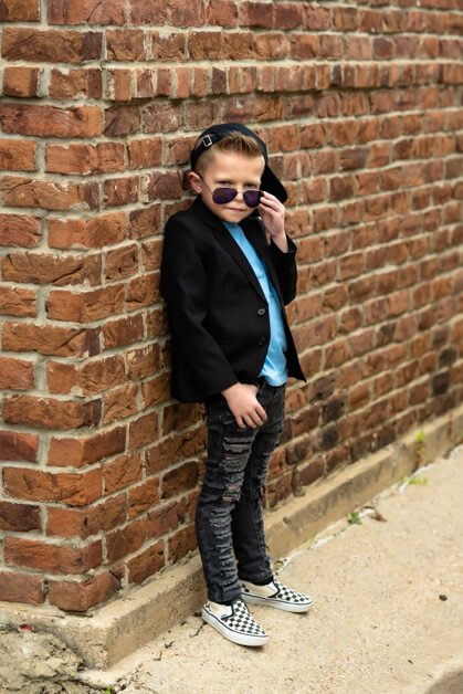 7-year-old boy leaning on a red brick building, dressed in ripped jeans, a light blue t-shirt, a black blazer, and checkered Vans sneakers pulling his sunglasses down to look over them during a mini photo session in downtown Roanoke, Texas.