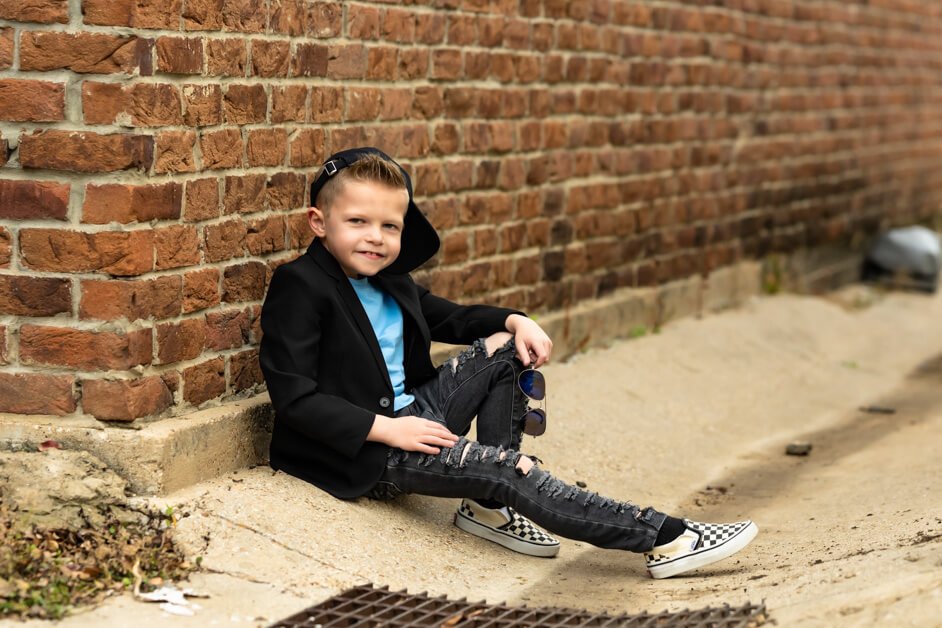 7-year-old stylish boy leaning casually against a red brick wall, dressed in ripped jeans, a light blue t-shirt, a black blazer, black ball cap and checkered Vans sneakers during a custom mini photo session in downtown Roanoke, Texas.