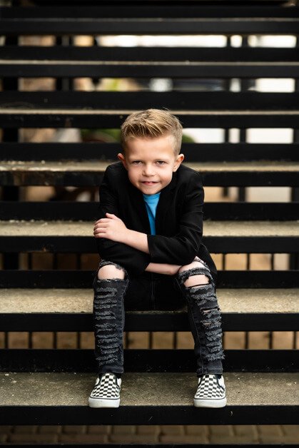 7-year-old boy sitting casually on a metal and concrete stairway outside a red brick building, dressed in ripped jeans, a light blue t-shirt, a black blazer, and checkered Vans sneakers during a mini photo session in downtown Roanoke, Texas.