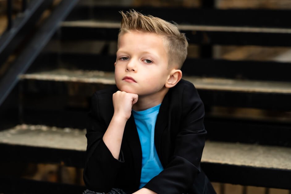 7-year-old boy leaning casually against a red metal bar in front of a brick wall, dressed in ripped jeans, a light blue t-shirt, a black blazer, and checkered Vans sneakers during a mini photo session in downtown Roanoke, Texas.