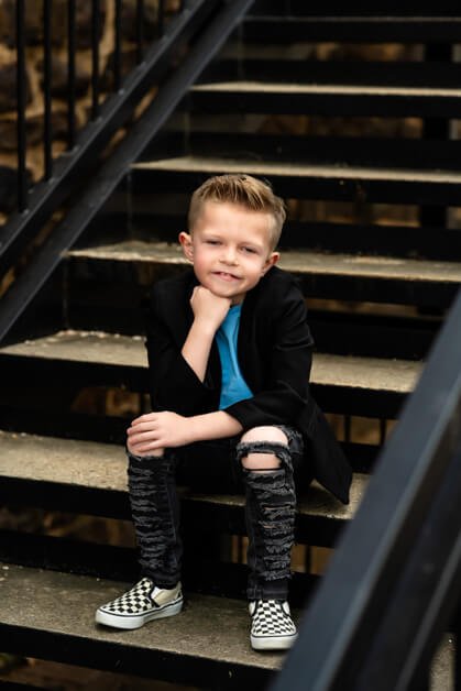 7-year-old boy sitting casually on a metal and concrete stairway outside a red brick building, dressed in ripped jeans, a light blue t-shirt, a black blazer, and checkered Vans sneakers during a mini photo session in downtown Roanoke, Texas.