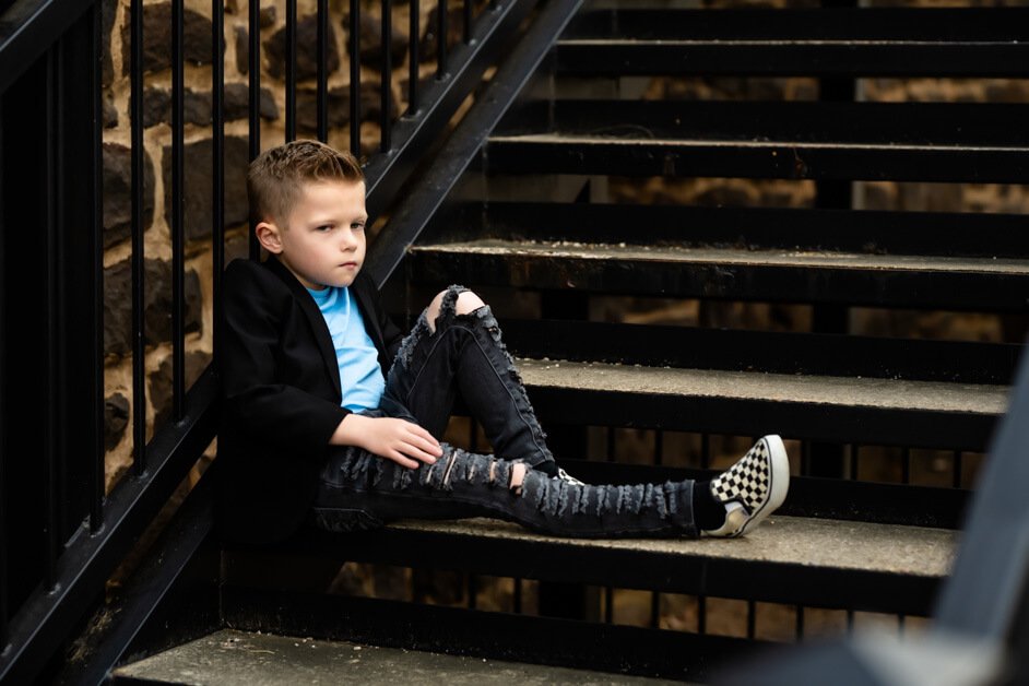 7-year-old boy sitting casually on a metal and concrete stairway outside a red brick building, dressed in ripped jeans, a light blue t-shirt, a black blazer, and checkered Vans sneakers during a mini photo session in downtown Roanoke, Texas.
