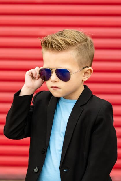 Close-up portrait of a 7-year-old boy adjusting his aviator sunglasses with a serious expression, dressed in a black blazer and light blue t-shirt, against a blurred red garage door in downtown Roanoke, Texas