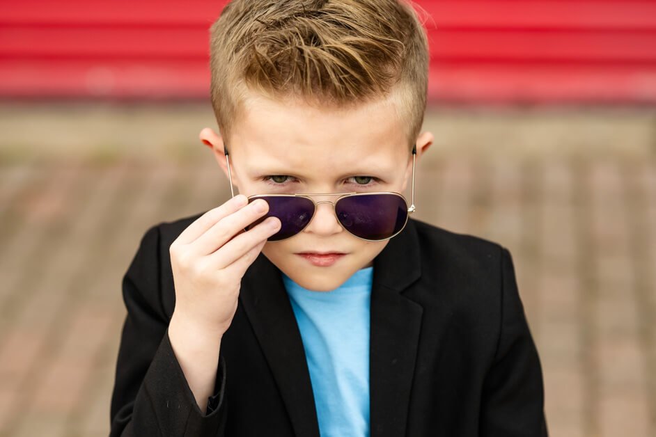 Close-up portrait of a 7-year-old boy adjusting his aviator sunglasses with a serious expression, dressed in a black blazer and light blue t-shirt, against a blurred red garage door in downtown Roanoke, Texas