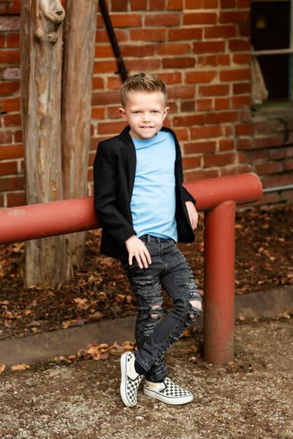 7-year-old boy leaning casually against a red metal bar in front of a brick wall, dressed in ripped jeans, a light blue t-shirt, a black blazer, and checkered Vans sneakers during a mini photo session in downtown Roanoke, Texas.